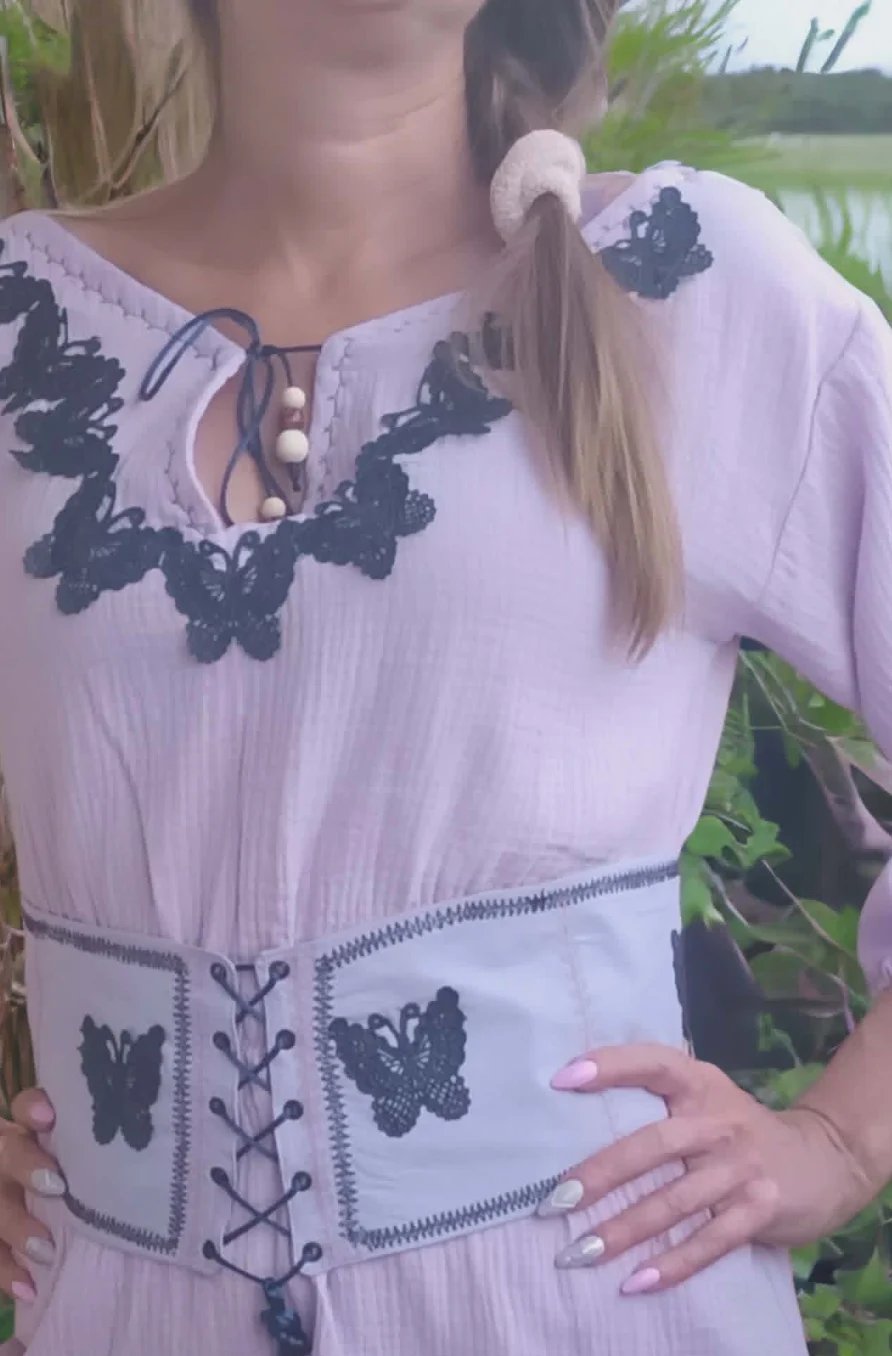 Woman wearing lavender dress with black butterfly embroidery and lace-up corset belt outdoors by water