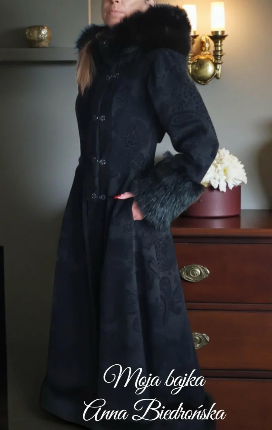 Woman wearing a black fur-trimmed coat with white fur cuffs, standing beside a wooden dresser with white flowers