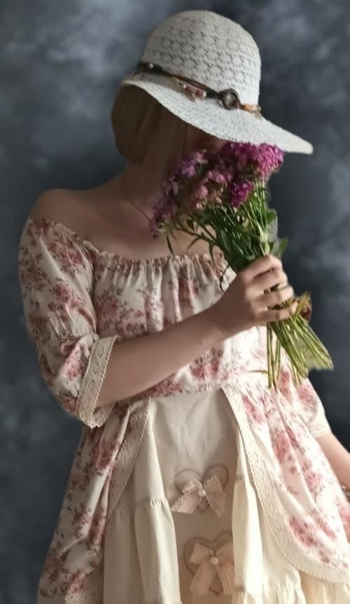 Person wearing a white straw hat and floral dress holding a bouquet of pink flowers against a blurred gray background