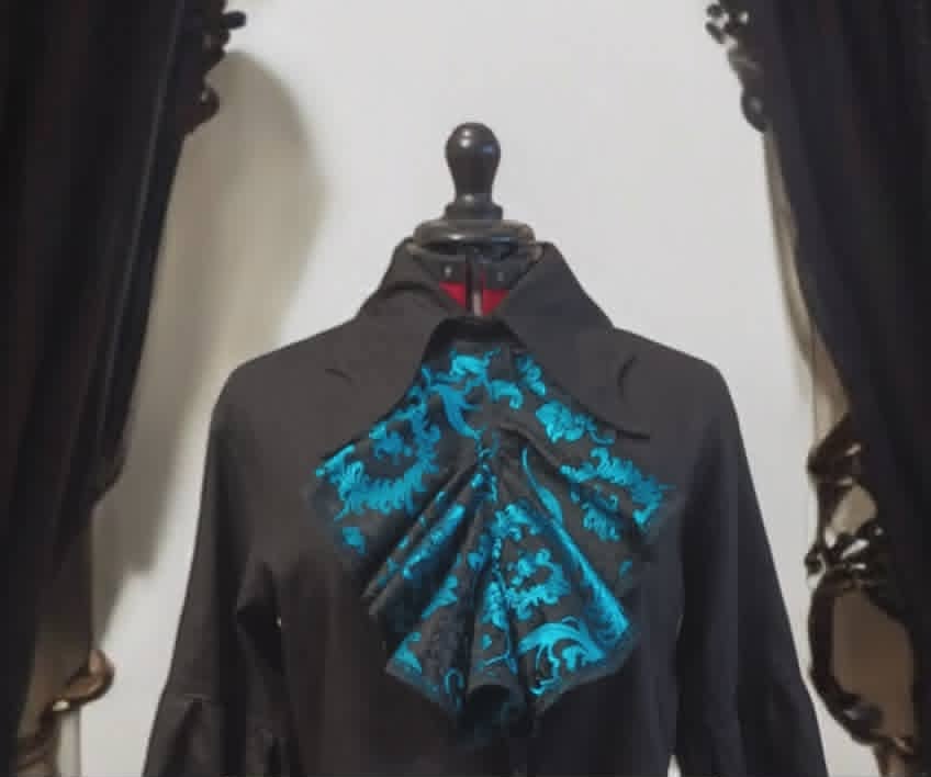 Black mannequin wearing dark robe with striking blue ornate patterned fabric draped across the back, displayed between dark curtains
