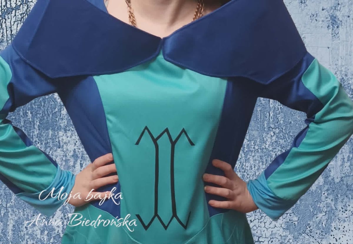 Person wearing a turquoise and navy blue tunic dress with a wide collar and mystical symbol on the chest