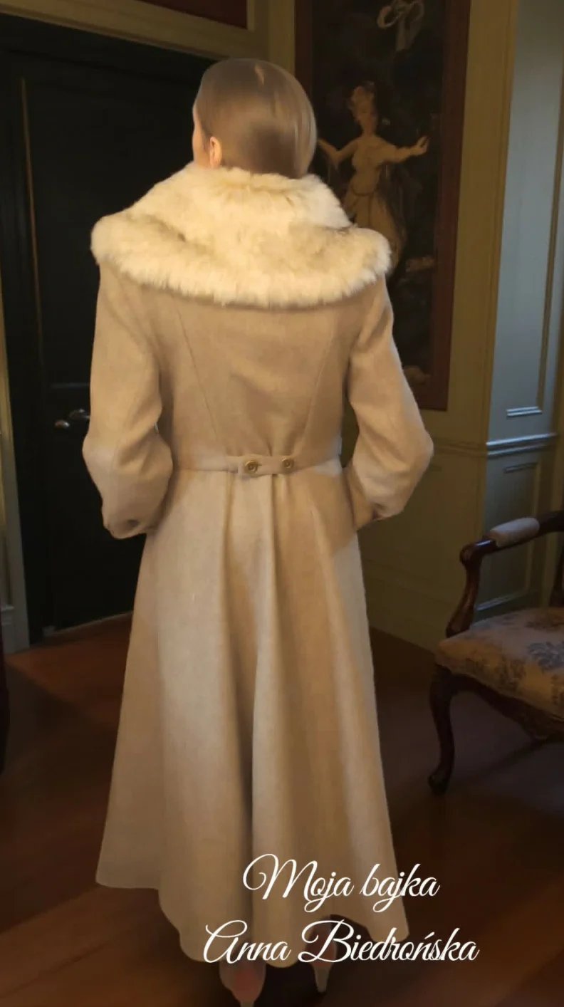 Woman wearing a beige coat with fur collar, viewed from behind in an elegant interior setting