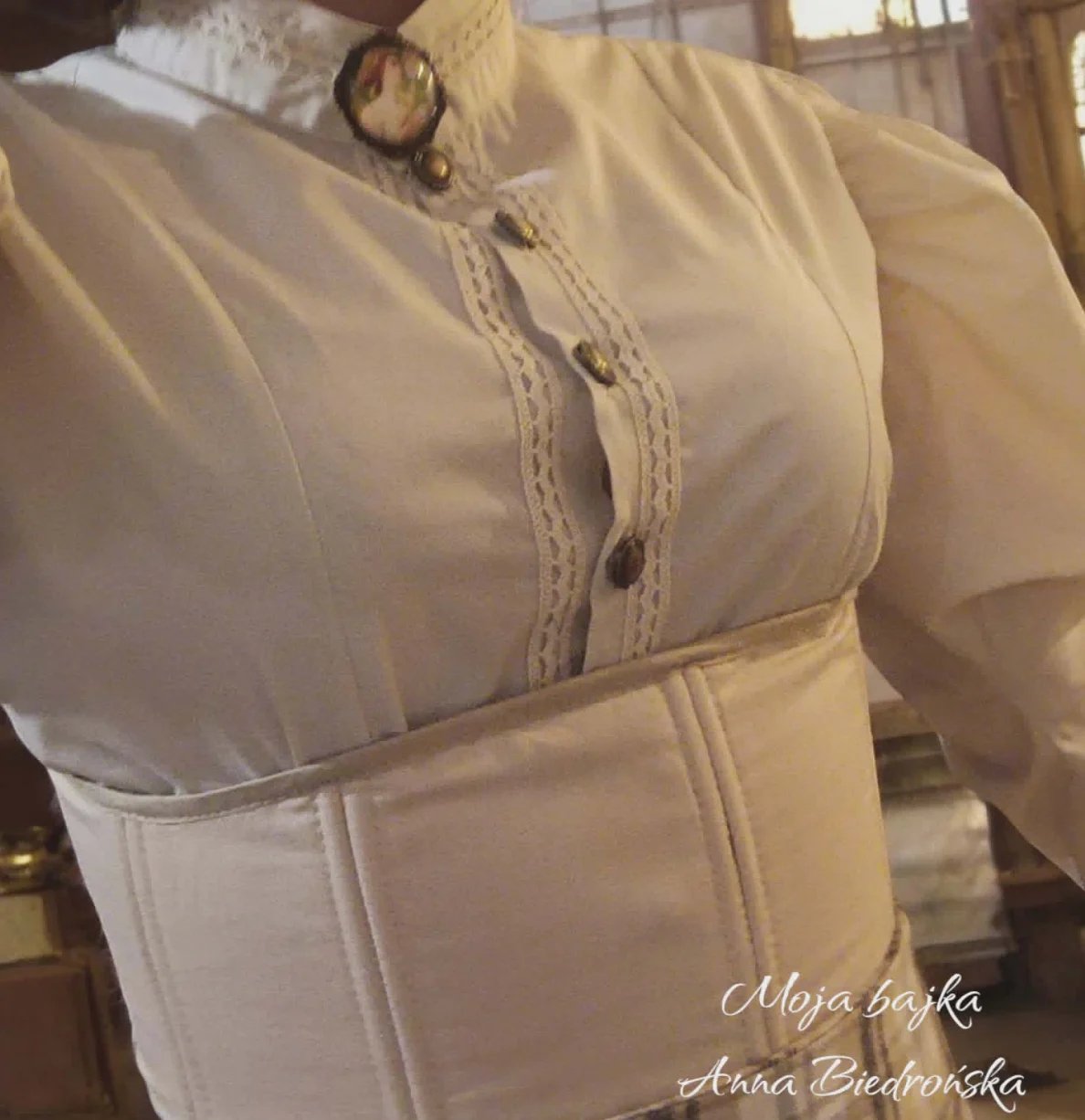Dress form displaying a beige corset with lace detailing and decorative buttons, shown on a mannequin indoors.