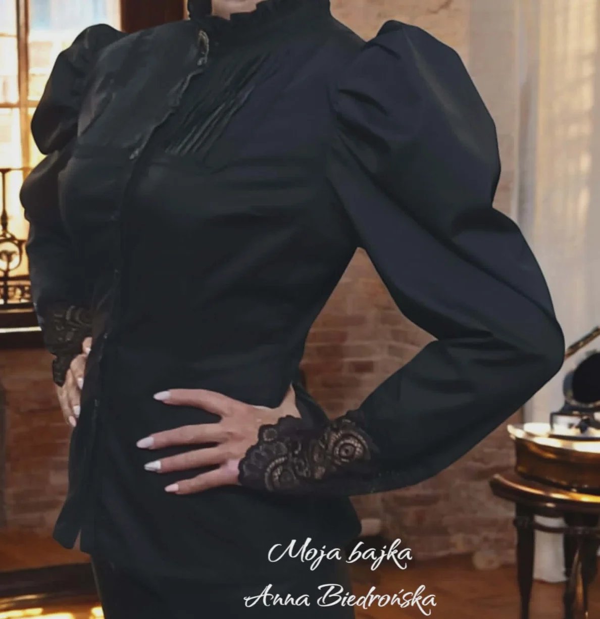 Woman wearing black blazer with puffed sleeves and lace cuff details, posing indoors with elegant home decor background