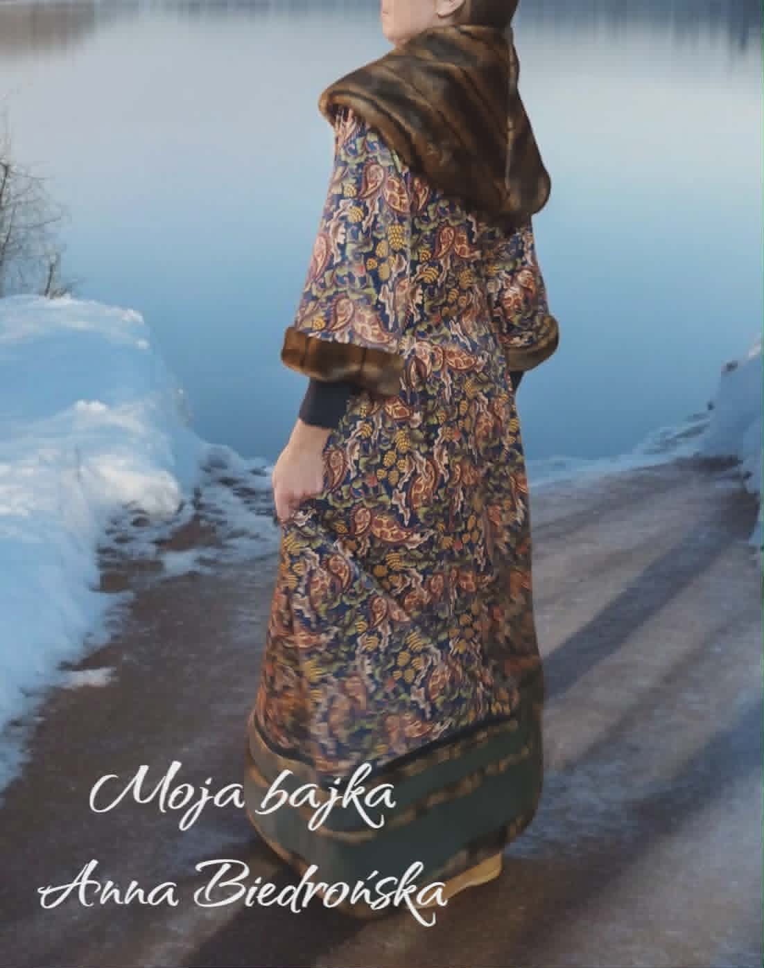 Woman in ornate patterned dress and brown fur stole standing by snowy lake shore, viewed from behind