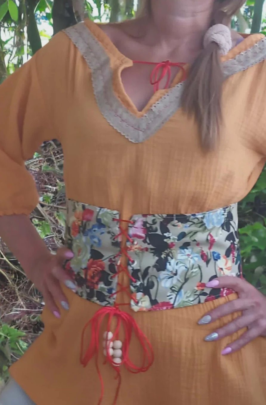 Woman wearing a mustard yellow dress with lace trim, floral corset belt, and red lacing details, standing outdoors with green foliage in background