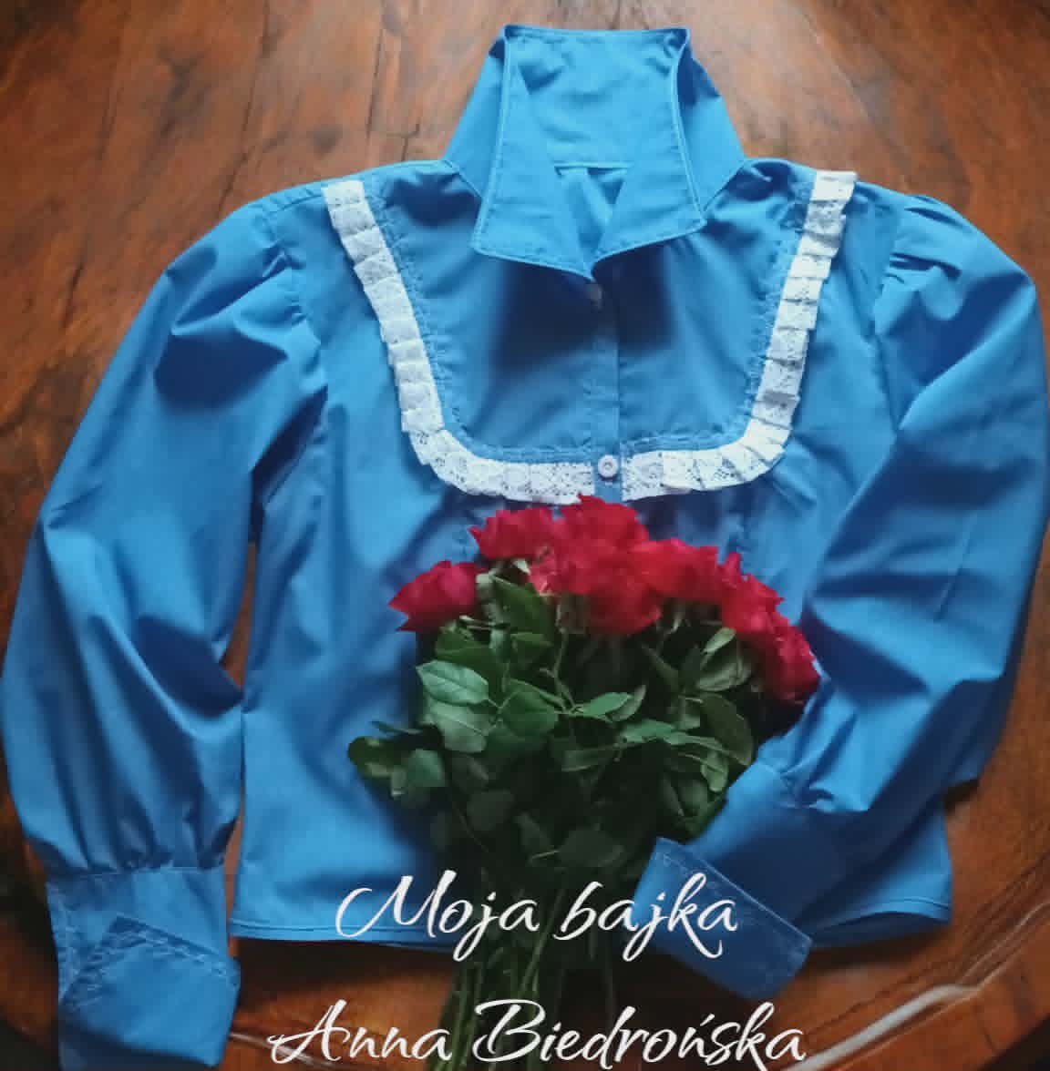 Blue traditional folk blouse with white lace trim and red roses on wooden background, with text Moja bajka and Anna Biedrońska