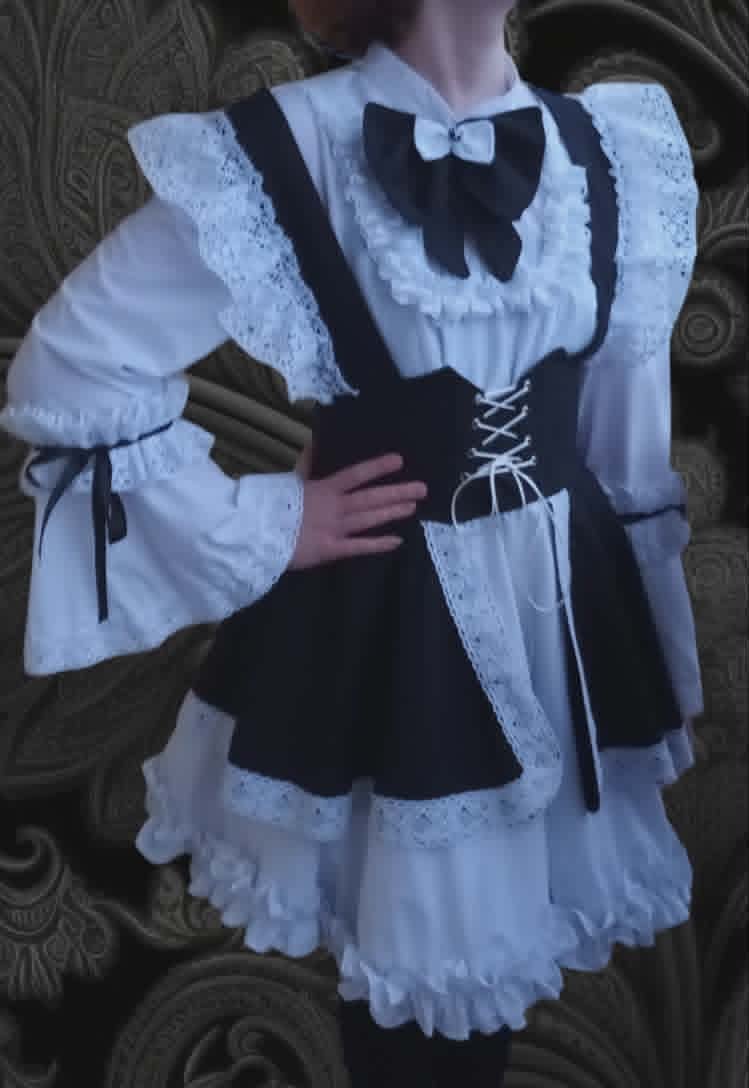 Person wearing Gothic Lolita fashion with white lace sleeves, black bow tie, black corset, and layered white frilled skirt against ornate gray wallpaper background