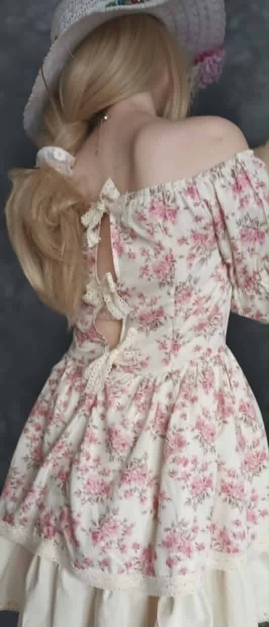 Porcelain doll wearing a white knit hat, off-shoulder floral dress with pink flowers, and cream lace trim, shown from behind against gray background