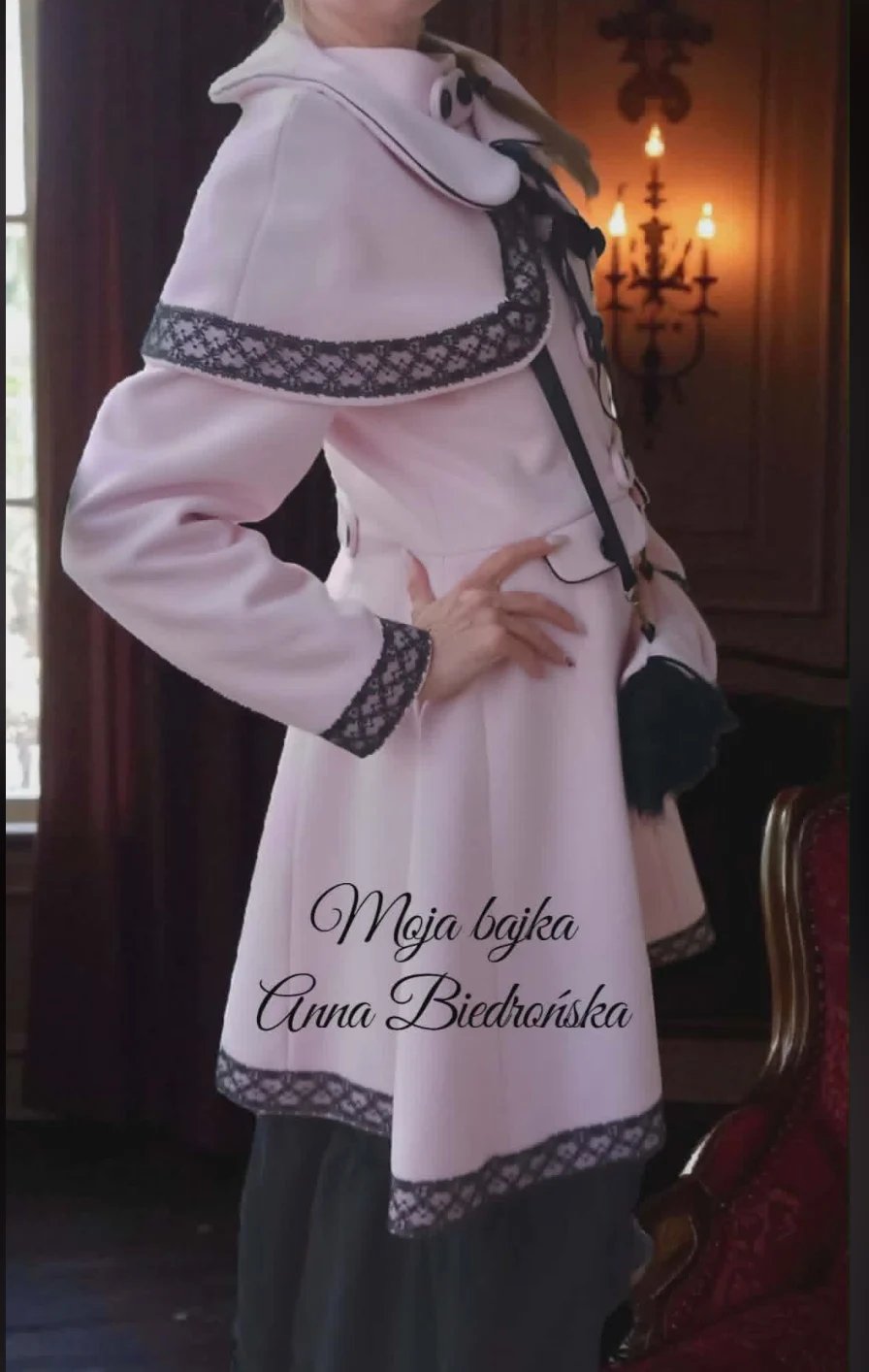 Person wearing a light pink coat with decorative geometric trim, standing indoors with a chandelier visible in the background