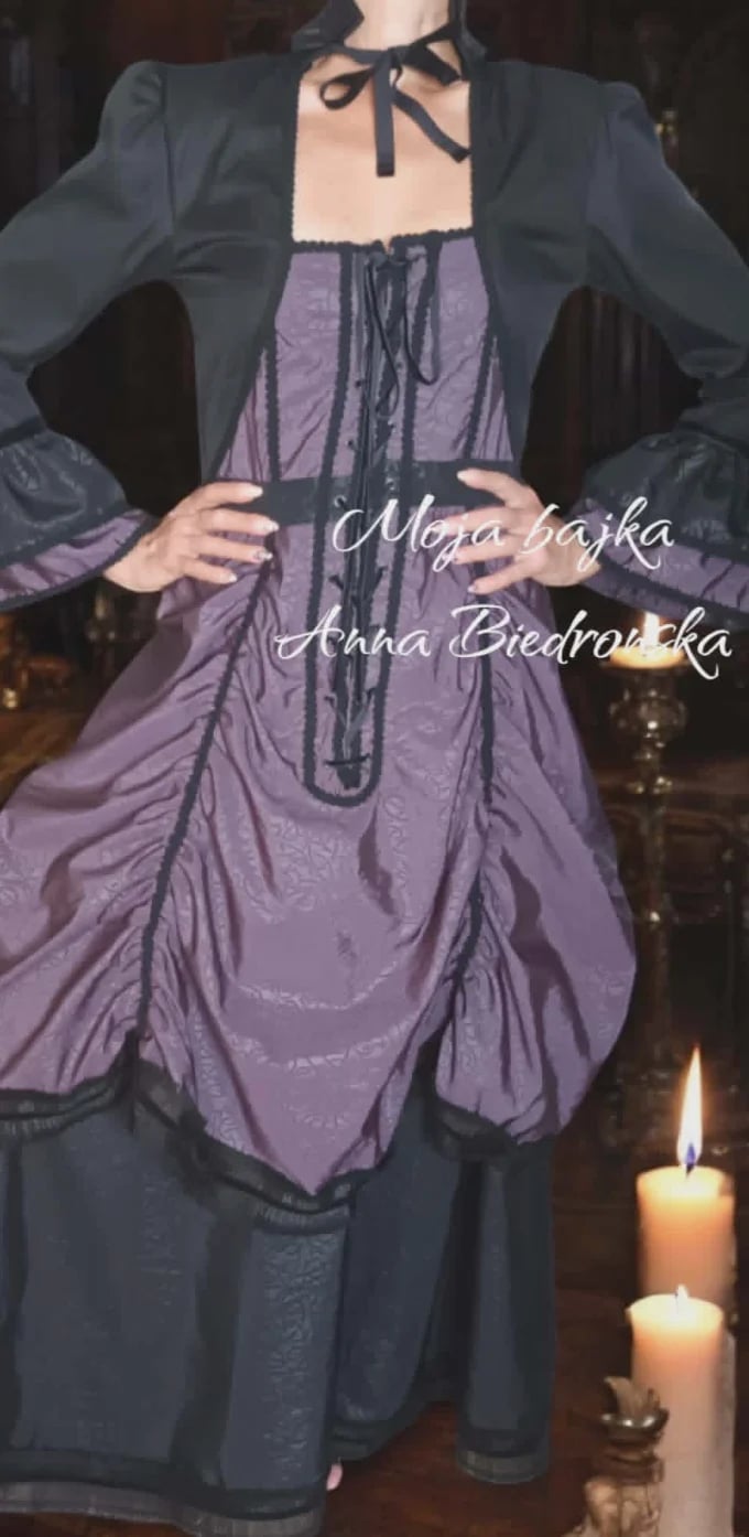 Person wearing gothic Victorian-style purple corset dress with black blazer and neck bow, standing with lit candles, text overlay with names
