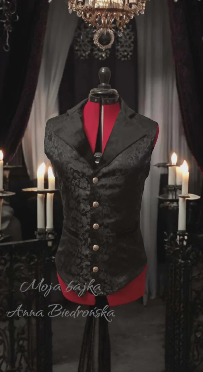 A dress form displaying a black ornate vest with red inner lining in a gothic-inspired room with candles and a crystal chandelier