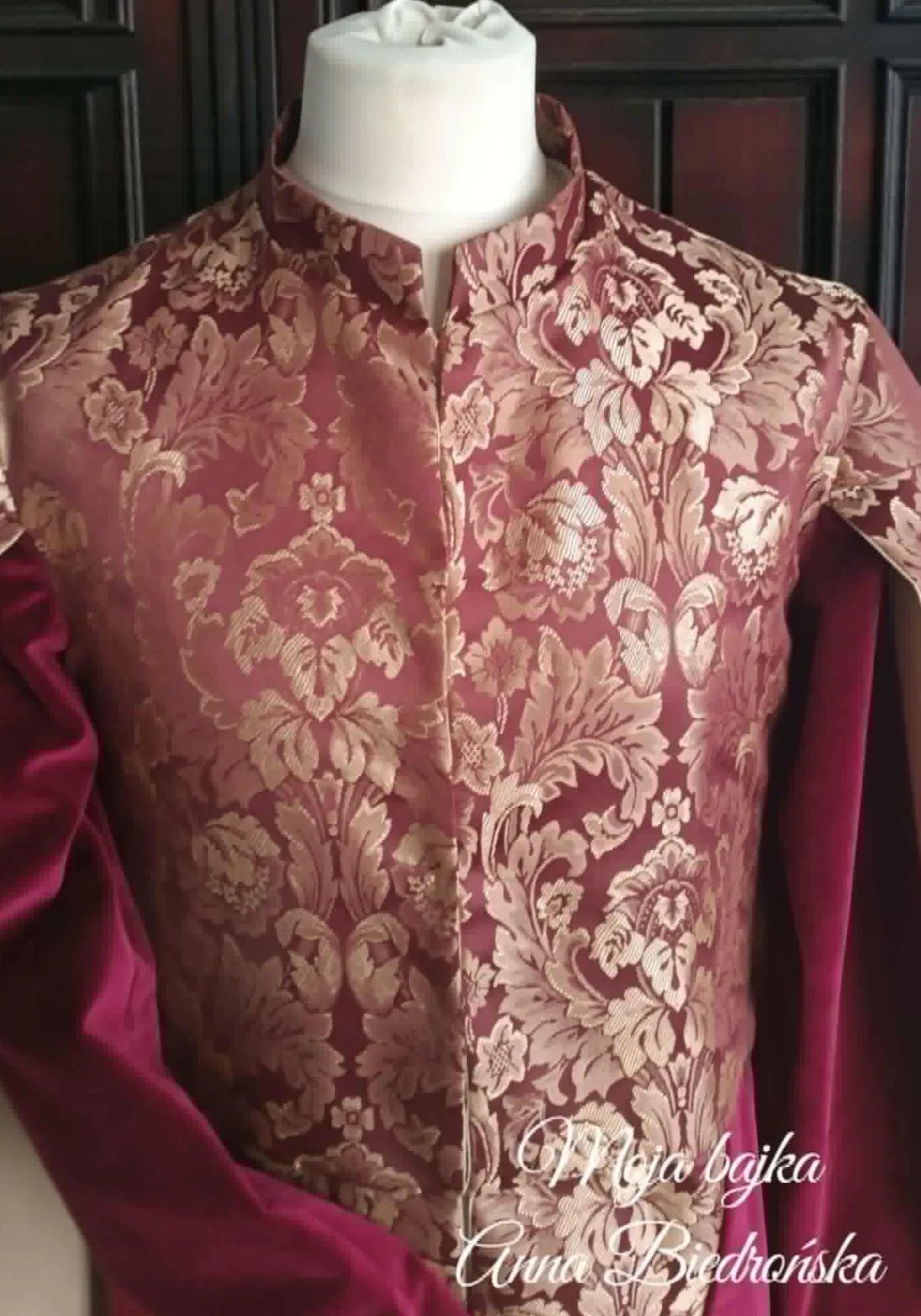 Burgundy ornate sherwani with gold damask embroidery on mannequin against dark paneled backdrop