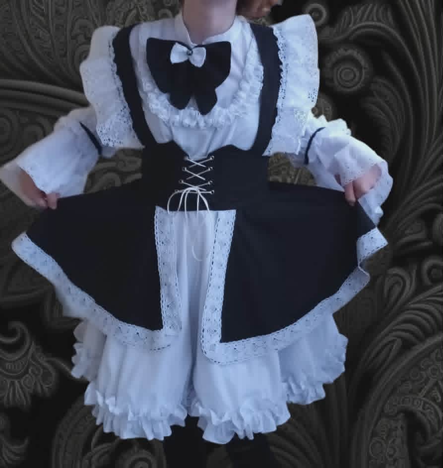 Child wearing a black and white lolita-style dress with ruffled petticoat, white lace collar, and black bow against patterned wallpaper background