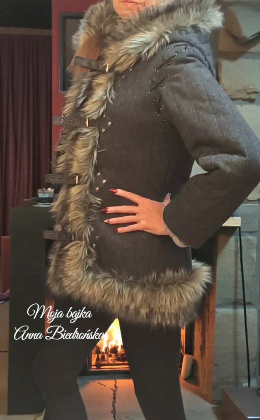 Person wearing black studded jacket with fur collar standing indoors with fireplace visible in background