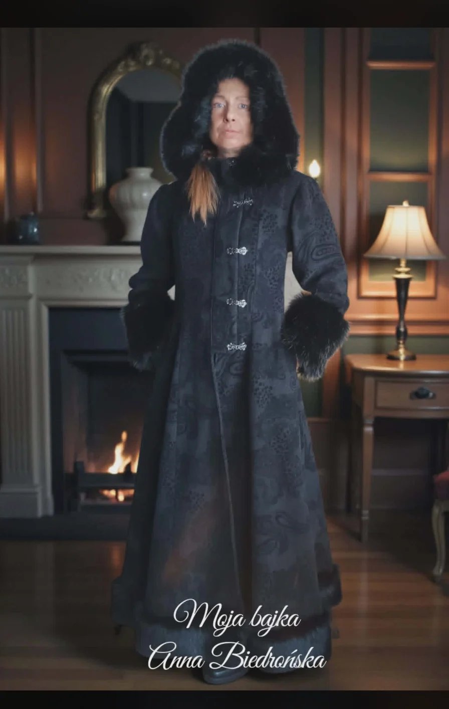 Woman in dark coat with fur trim standing in elegant living room with fireplace and warm lighting