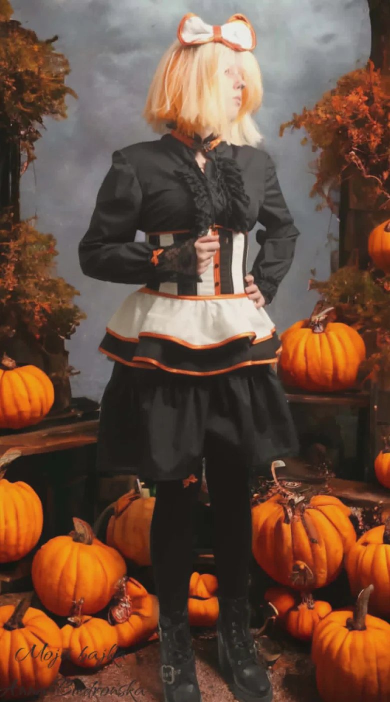 Person in autumn-themed costume with blonde hair, orange bow, black jacket and layered skirt standing among pumpkins against fall backdrop