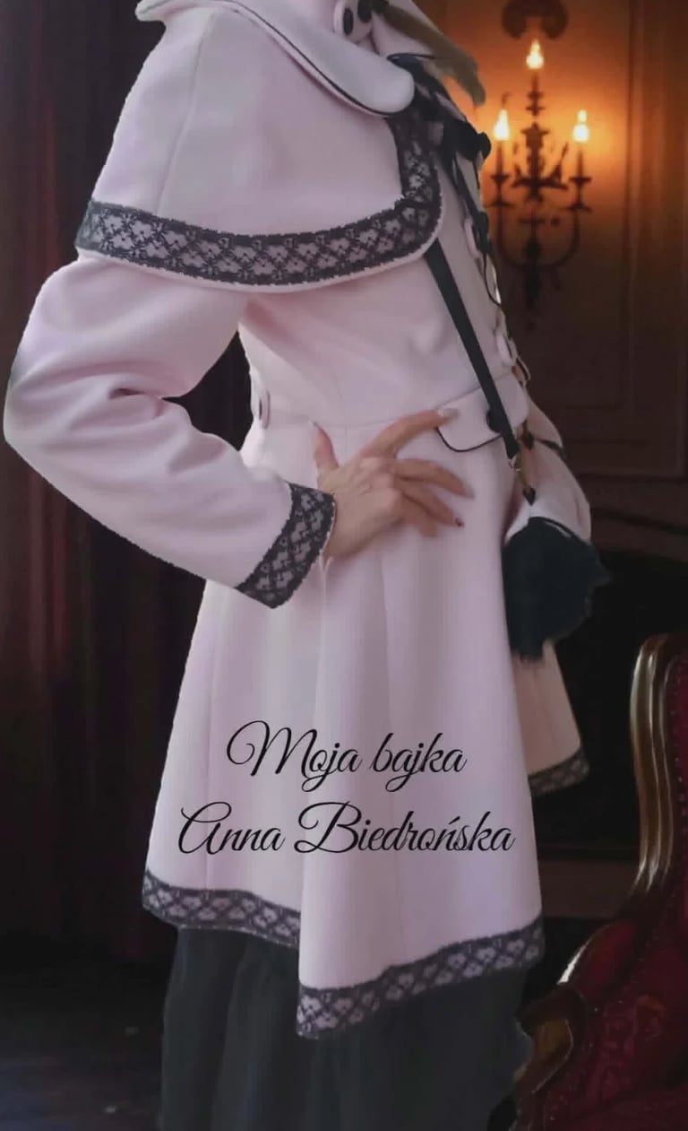 Woman wearing traditional Polish white coat dress with black decorative trim and embroidered belt, displaying text on the garment, standing indoors with ornate chandelier in background
