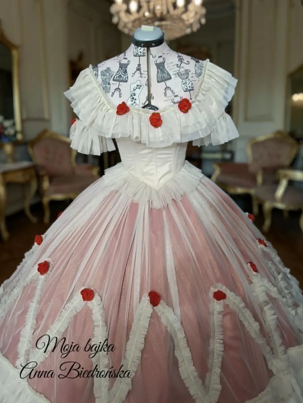 Elegant gown with white ruffled off-shoulder neckline, printed bodice detail, and pink tulle skirt adorned with red floral embellishments
