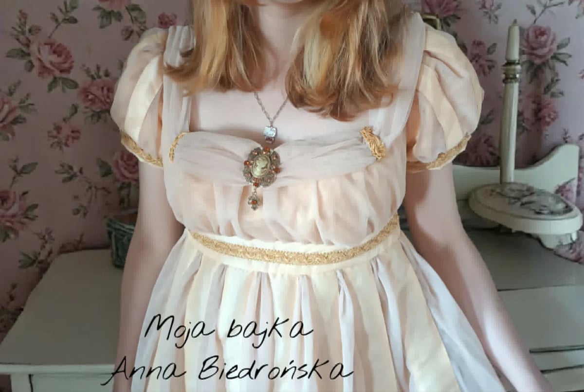 Young woman wearing a cream-colored romantic dress with puffed sleeves and ornate necklace, standing against floral wallpaper