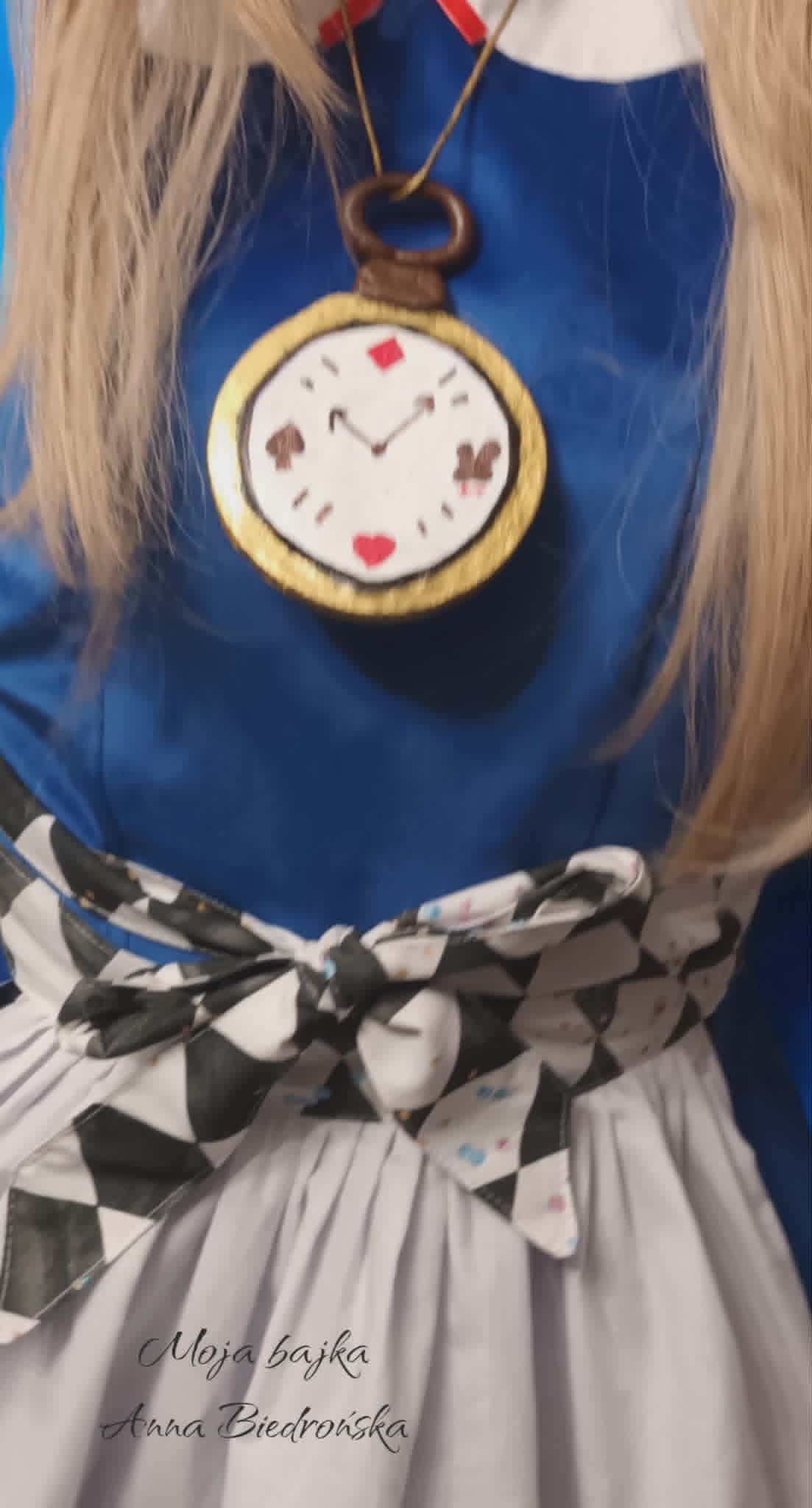 Character in blue shirt wearing a gold-framed pocket watch pendant with playing card suit symbols on a brown cord