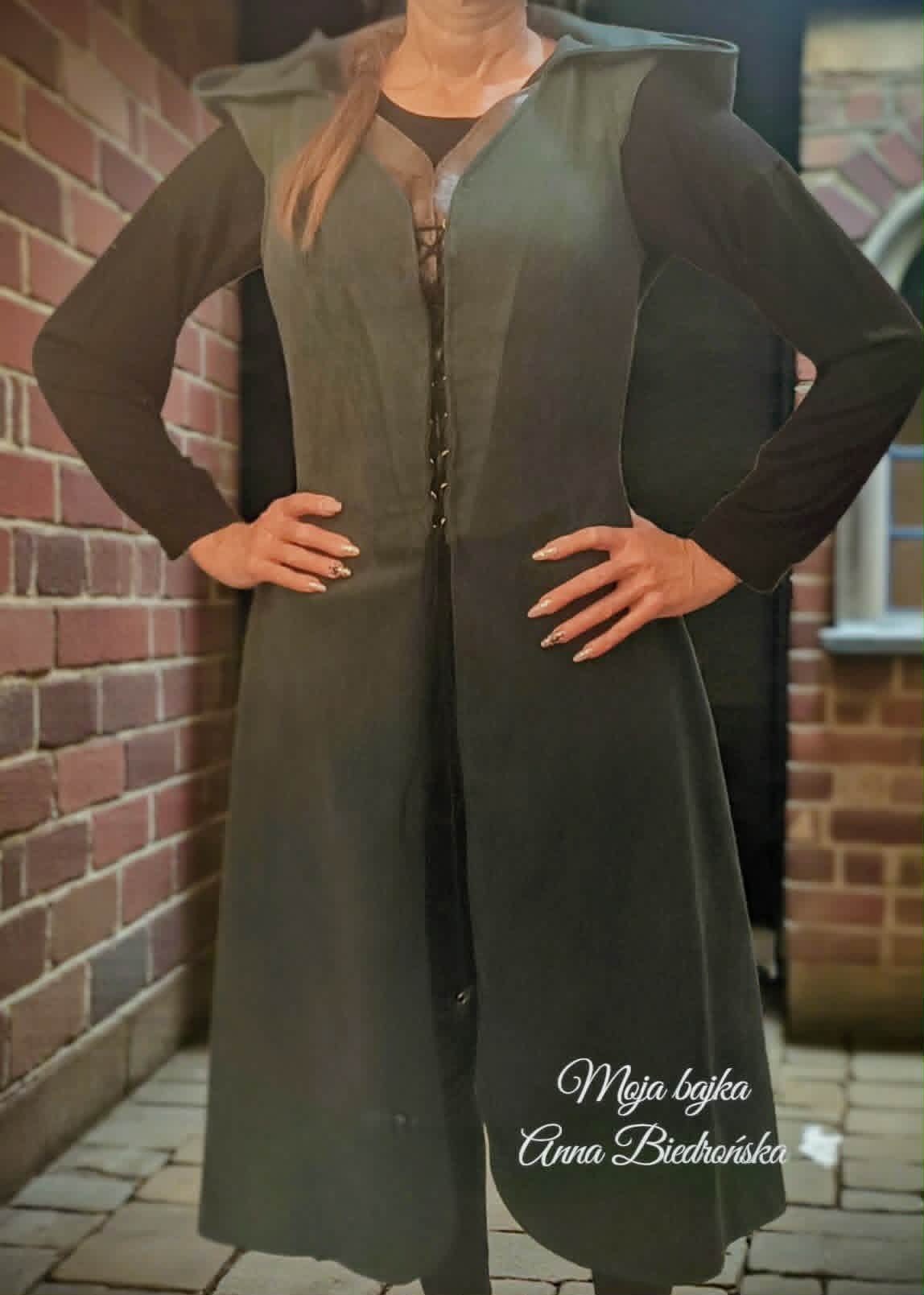 Woman in dark olive long cardigan with black long sleeves standing against brick wall with hands on hips