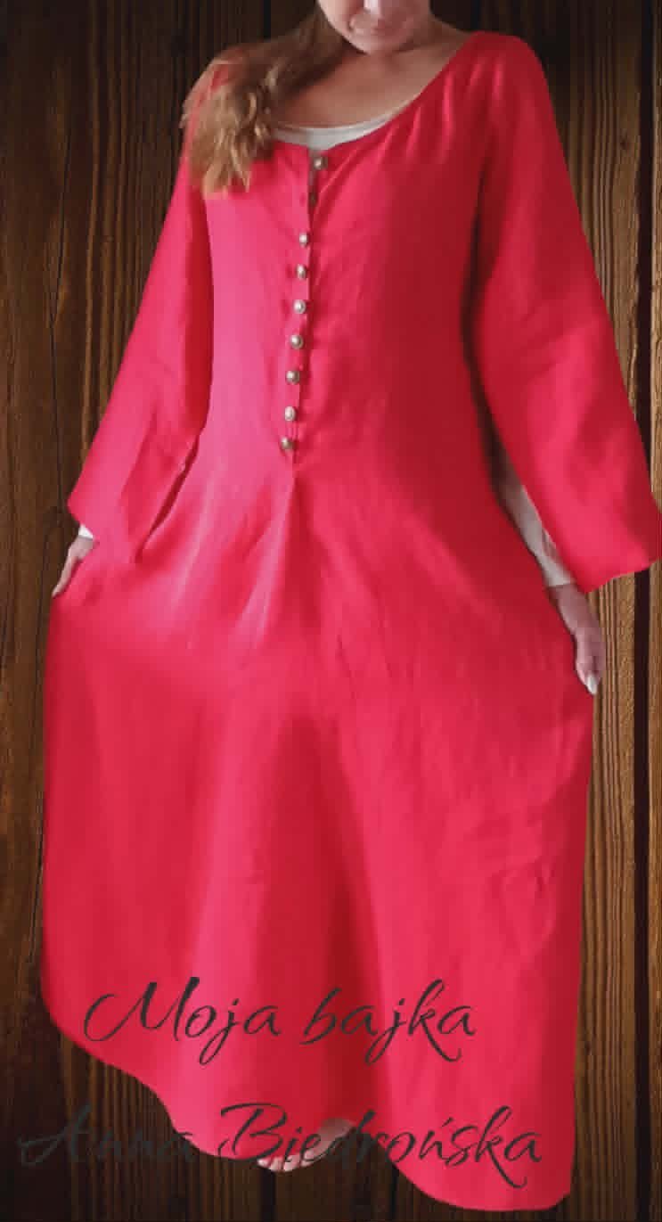 Woman wearing a bright pink long-sleeved dress with pearl button details and white trim, standing against a wood background