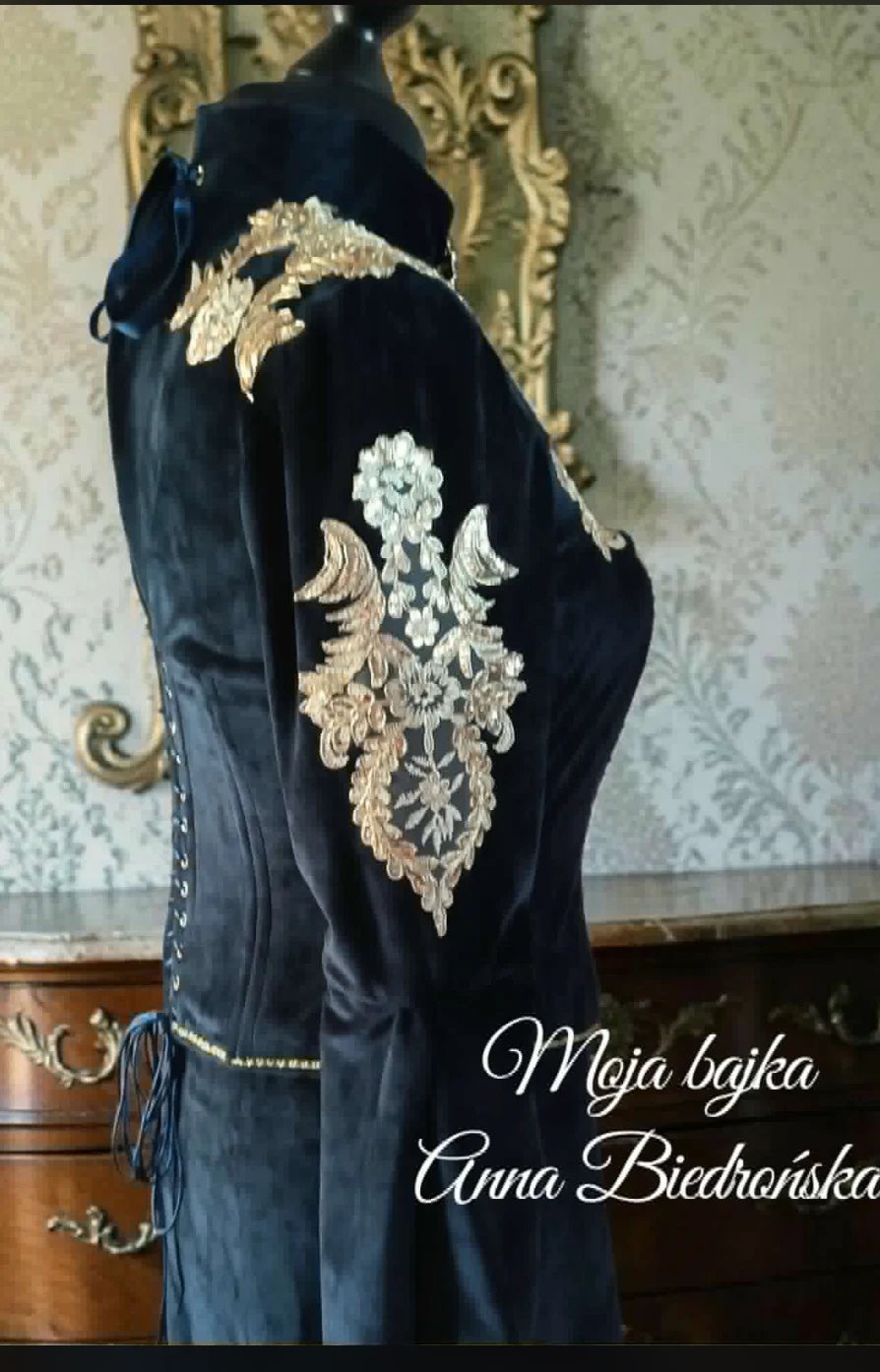 Black formal robe with ornate gold and white embroidered details on the back, displayed on a dress form against decorative wallpaper and gilded frame