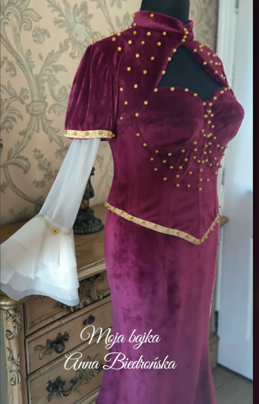 Deep purple velvet gown with gold beaded detailing on a dress form, featuring white draping fabric and ornate damask wallpaper background