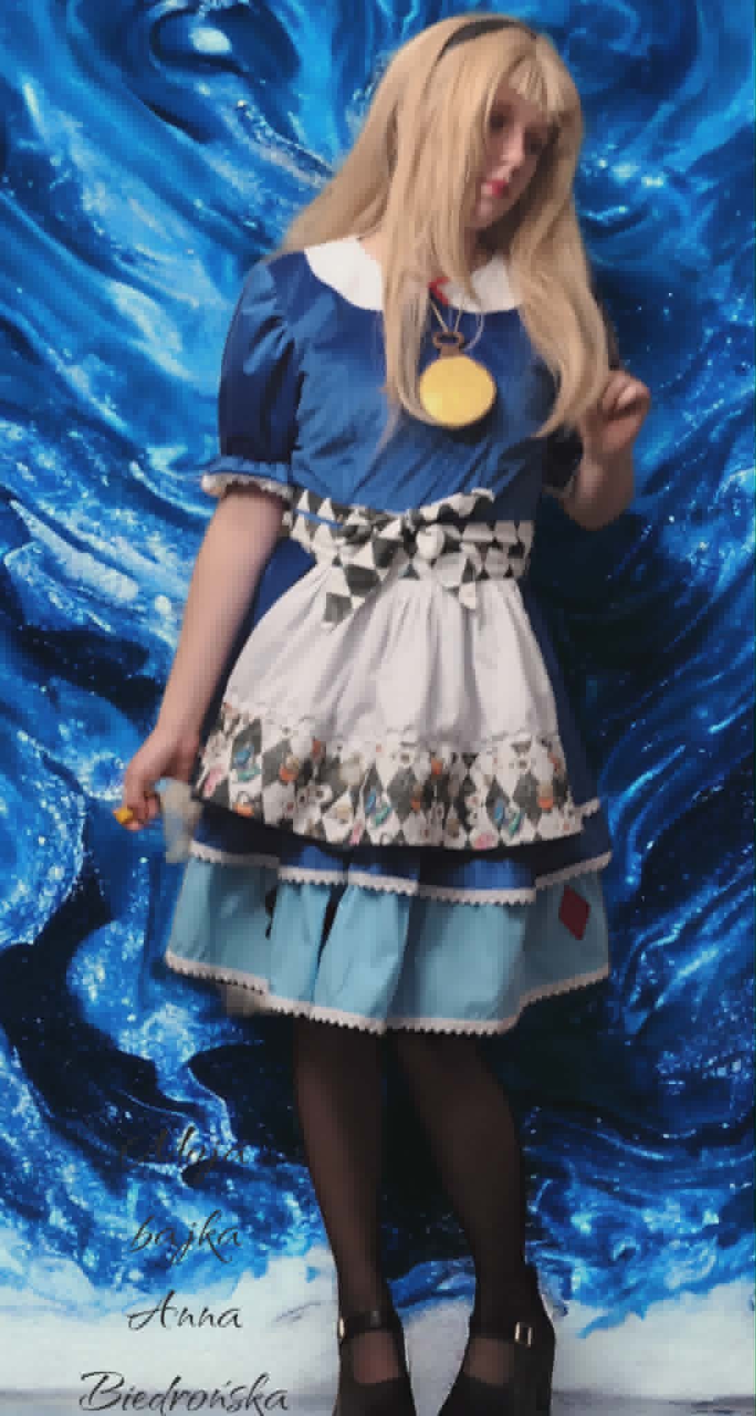 Person in blonde wig wearing blue and white anime-style dress with yellow pendant against blue swirling background