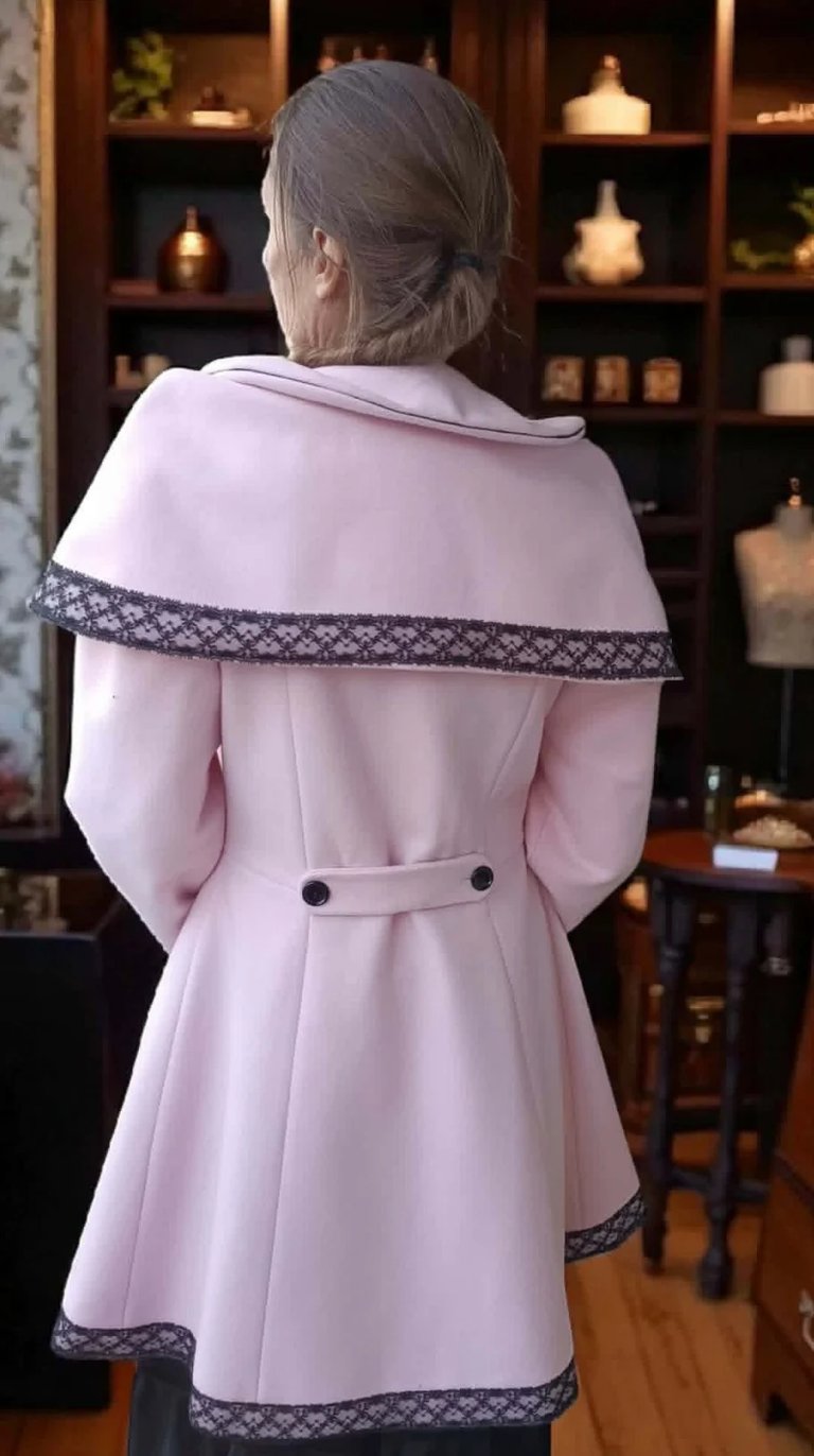 Elderly woman from behind wearing pink coat with patterned trim and detachable cape collar in an upscale interior with shelving
