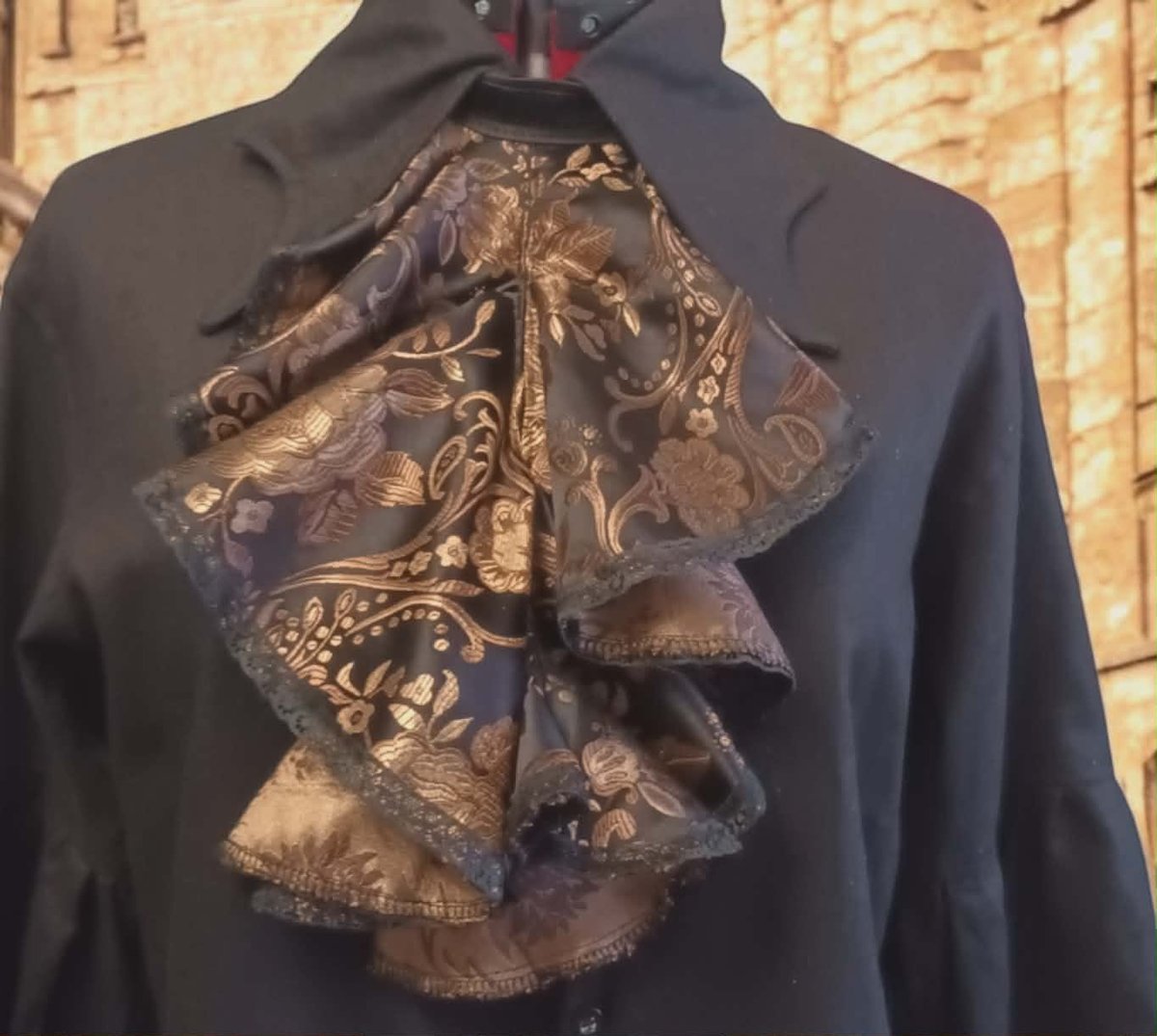 Black blazer with bronze patterned necktie or bow detail displayed on a hanger against a brick wall background
