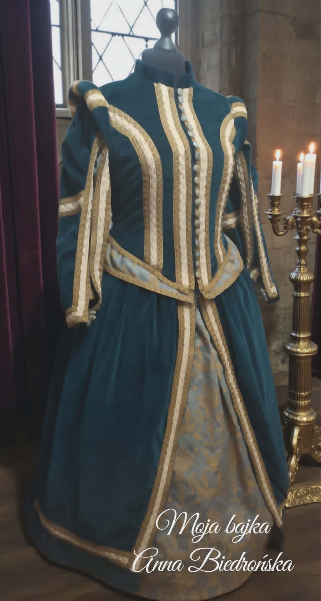 Blue medieval dress on mannequin with gold trim, displayed indoors with candlabra and window in background