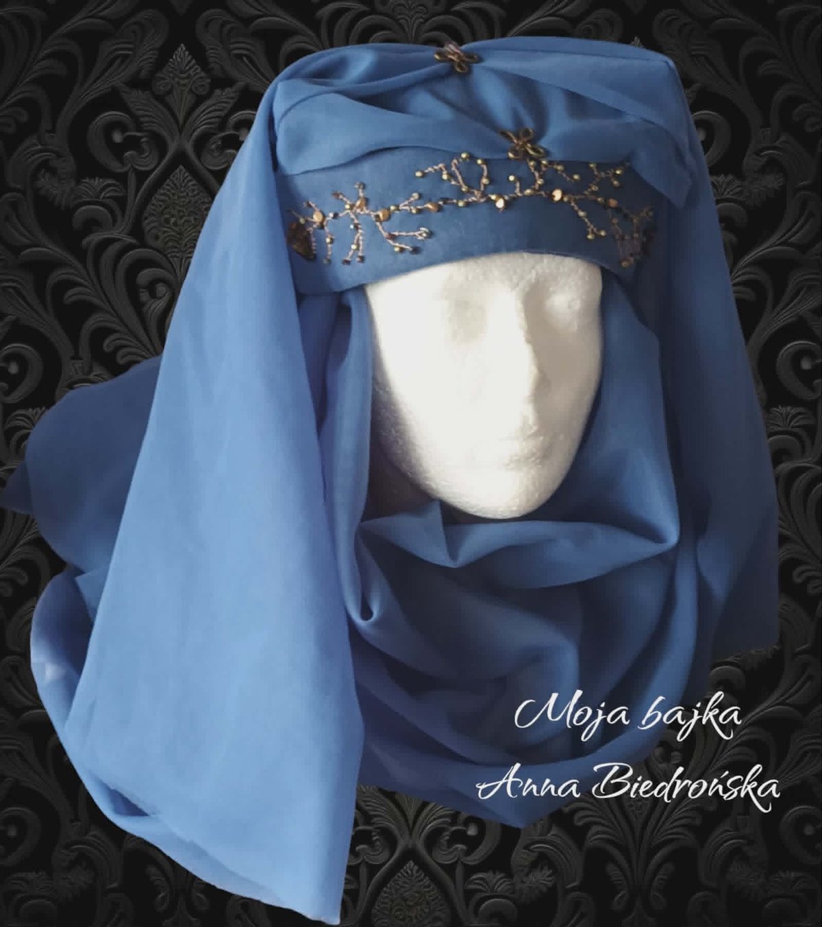 White mannequin head wearing blue headscarf with gold beaded headband against ornate dark damask background