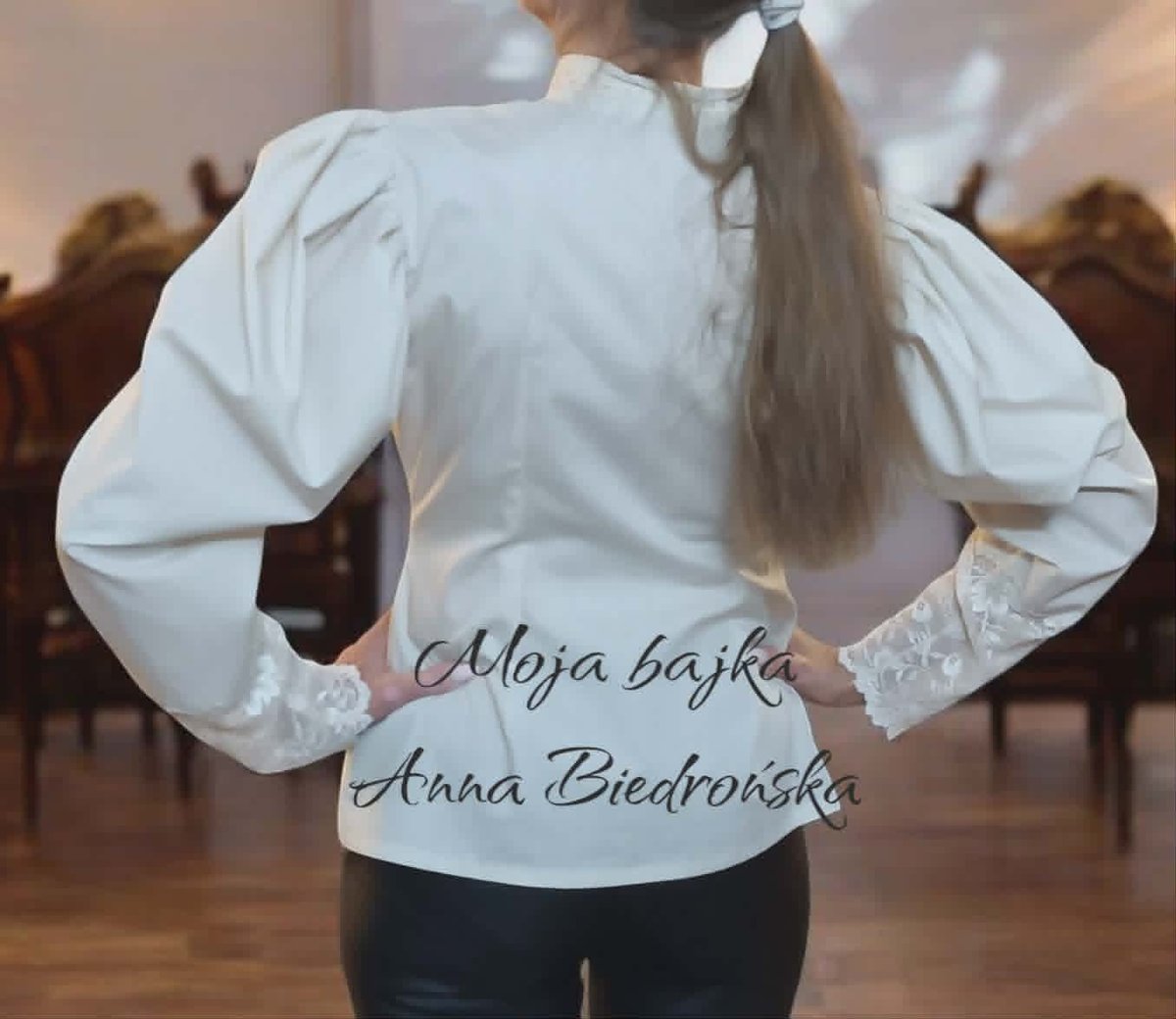 Person wearing white puff-sleeved blouse with text on back, shown from behind with long hair in ponytail