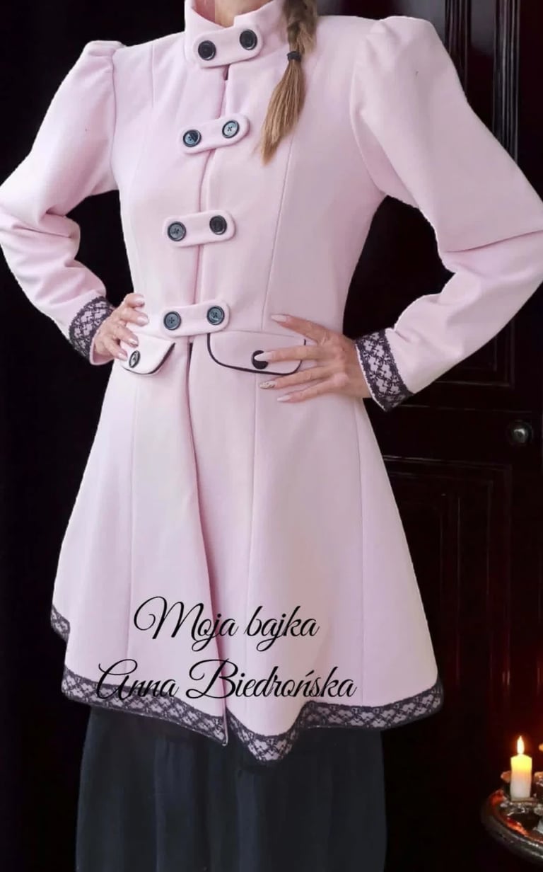 Woman wearing a light pink vintage-style coat with puffed sleeves, double-breasted buttons, and black lace trim over a dark skirt