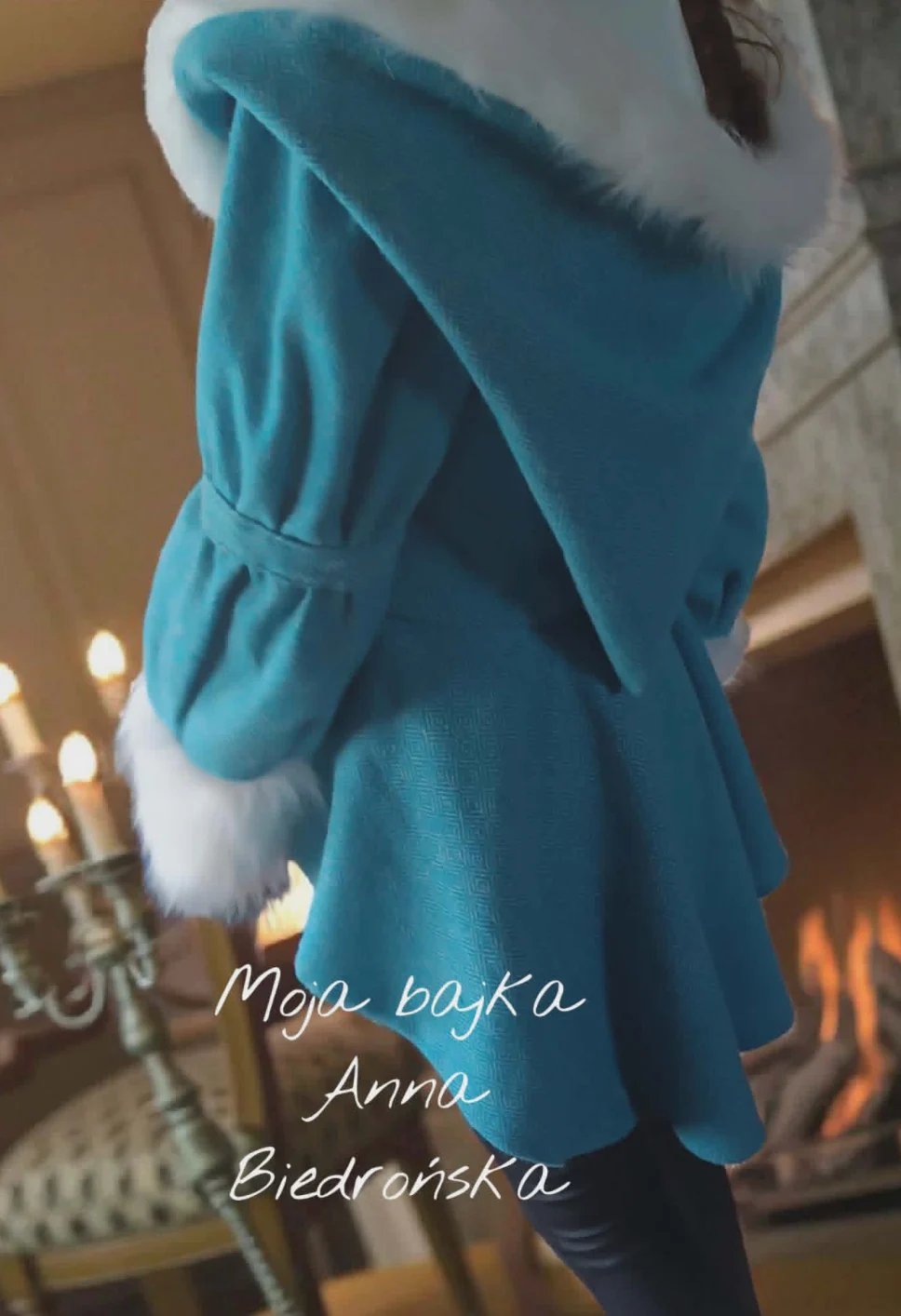 Woman wearing a turquoise blue coat with white fur-lined hood and collar in an elegant interior setting