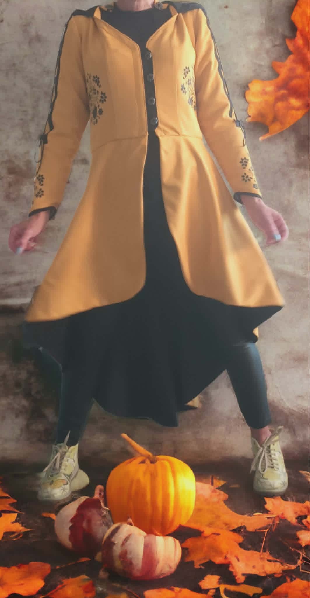 Person wearing a yellow cardigan over a black dress standing behind autumn leaves and small pumpkins