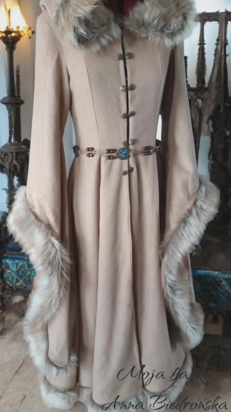 Cream-colored medieval-style dress with fur trim at collar, cuffs, and hem, decorated with ornate belt and toggles, displayed on mannequin in antique setting