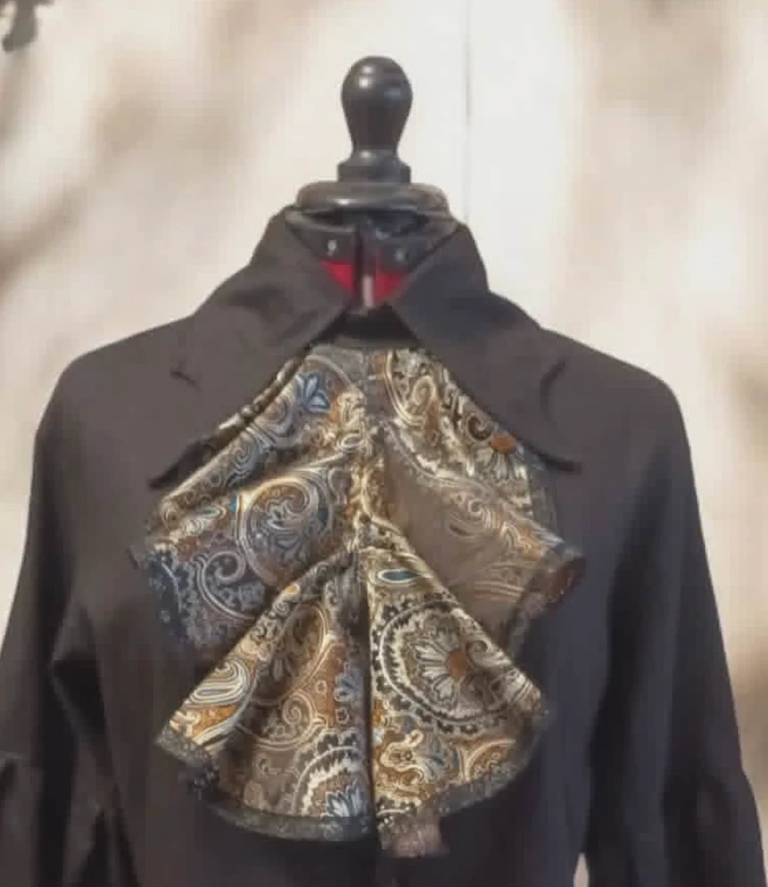 Black mannequin wearing a black long-sleeved garment with a gold and silver ornate paisley bow detail and red tie at the neckline