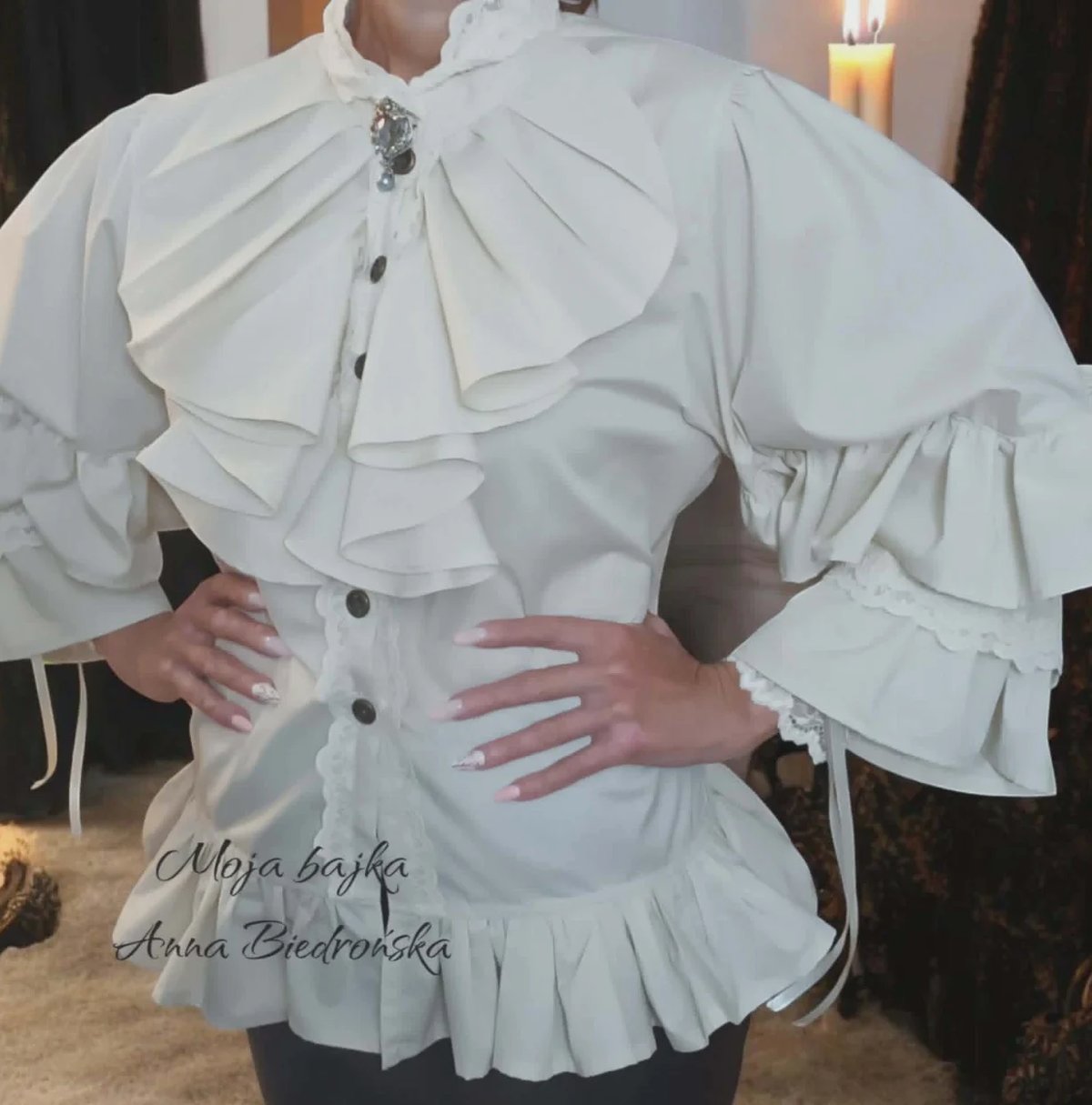Person wearing a white ruffled blouse with dramatic collar and puffed sleeves, hands on waist, posed indoors