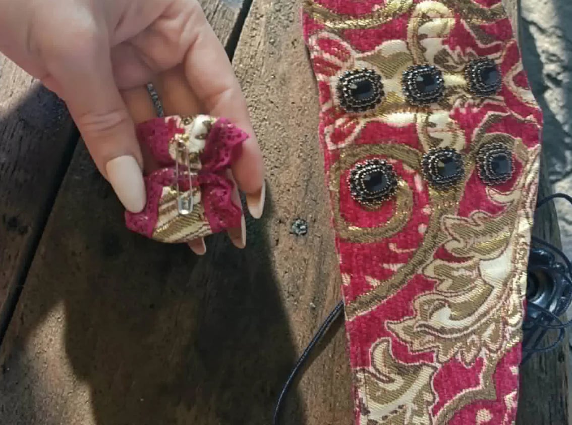 Hand holding a decorative brooch next to an ornate pink and gold embroidered fabric with jeweled details