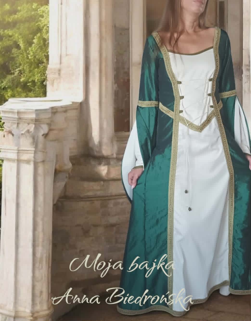 Woman wearing medieval-style green velvet overdress with gold trim over white linen gown, standing in stone archway