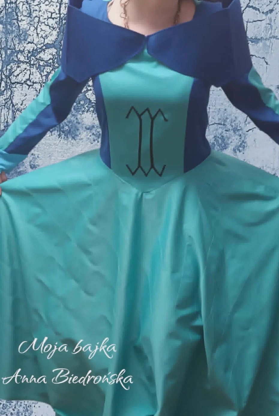Person wearing turquoise and navy dress with Scorpio symbol on chest, against weathered wall background