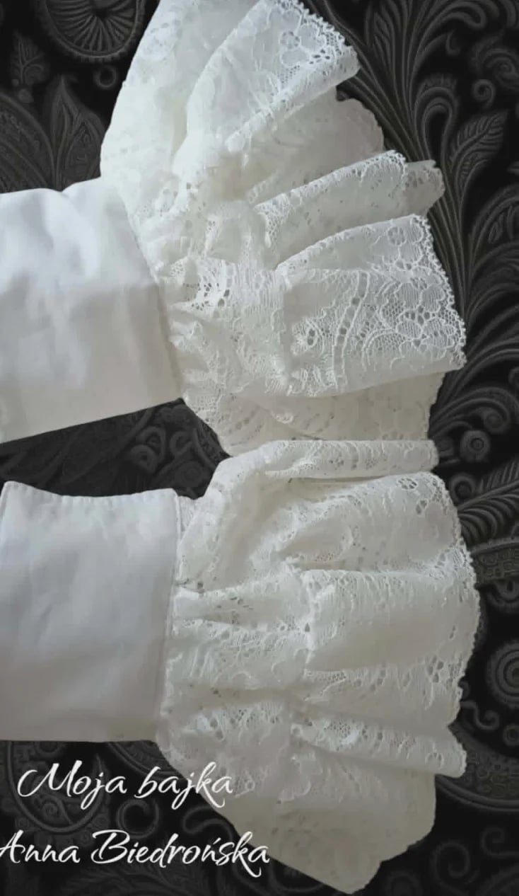 Two hands wearing delicate white lace gloves with ruffled cuffs against a dark patterned background
