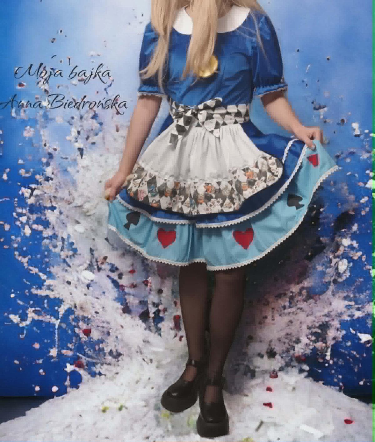 Woman in Alice in Wonderland-inspired costume with blue blouse, white apron with heart details, and black tights against colorful paint splatter background