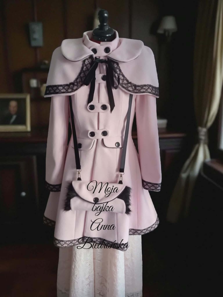 Pink dress on mannequin with black lace collar, white buttons, and text reading Meia Bajka and Anna Bialosuka displayed indoors