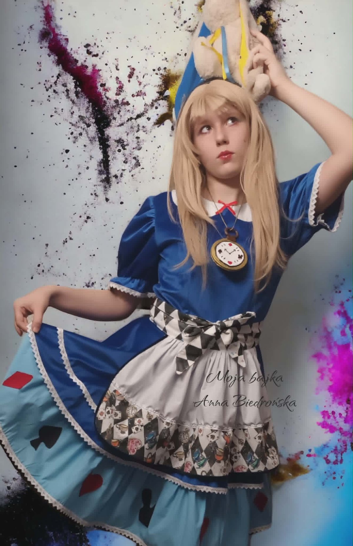 Cosplayer dressed as anime character in blue and white outfit with blonde wig and crown, surrounded by colorful paint splashes