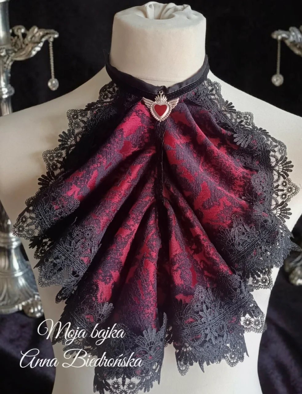 Decorative fabric bow brooch featuring deep red velvet center with black lace trim and heart-shaped pendant, displayed on dress form