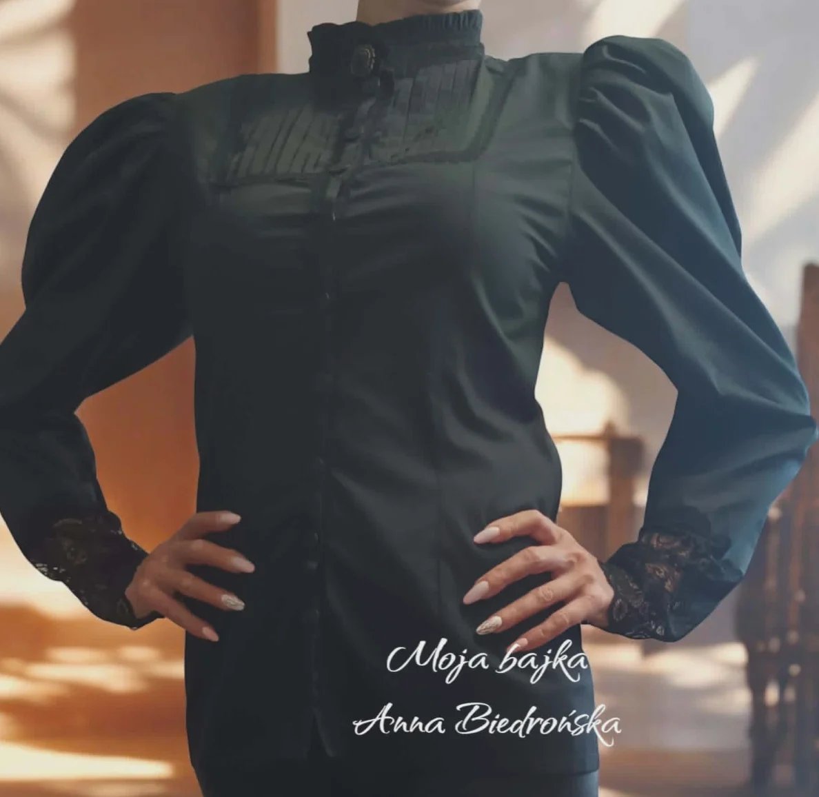 Woman wearing black puffed sleeve blouse with lace cuffs, hands on hips, sunlit interior background